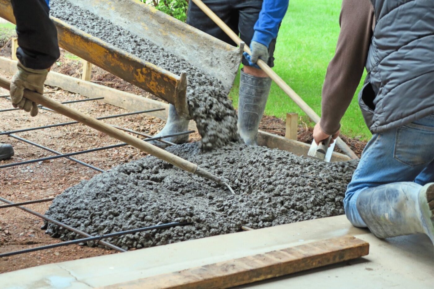 Three workers from Naples Concrete Solutions are pouring and spreading concrete over a wooden and metal framework using shovels and rakes. One worker, wearing rubber boots and a blue jacket, assists as the gray, wet concrete flows from a chute. The ground and grass in Naples, FL can be seen nearby.