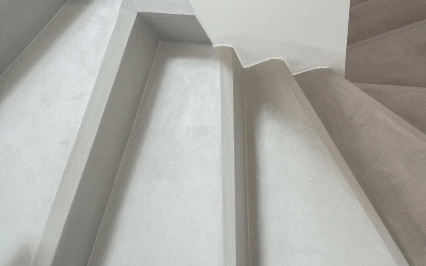 A top-down view of a modern staircase with light gray concrete steps and sharp, angled edges. The image captures the corner of the stairwell where the steps change direction, demonstrating clean lines and minimalist design.