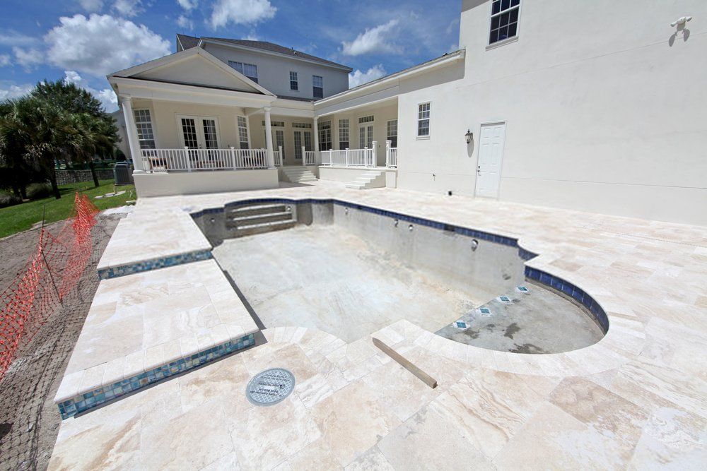 A large in-ground swimming pool under construction is set in a spacious backyard with a beige stone patio and a decorative pool deck. The pool is empty, and its edges are lined with blue tiles. A two-story house with a wraparound porch and white exterior is in the background. Contact Naples Concrete Solutions for a free estimate.