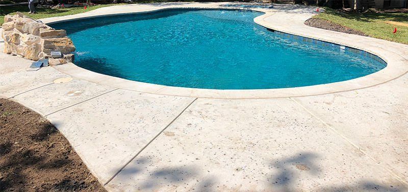 A curved swimming pool with clear blue water is surrounded by a wide, light-colored decorative pool deck from Naples Concrete Solutions. There is a small stone structure on the left side near the pool's edge, possibly a water feature or decorative element. The landscape includes grass and shrubs. Contact us for a free estimate today!