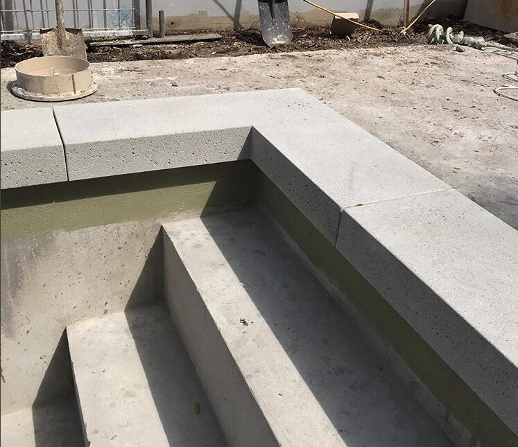 A partially constructed pool with concrete steps is shown, courtesy of Naples Concrete Solutions. The surrounding area appears to be under construction, with materials and debris visible in the background. The pool's interior is unfinished, and a railing just beyond the edge hints at a future decorative pool deck. For a free estimate, contact us today!