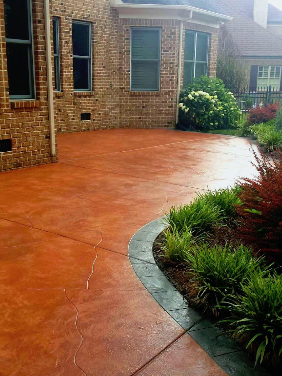 A red-toned decorative concrete patio curves around lush green landscaping and touches a brick house with large windows. The garden bed features a variety of plants, including grasses and blooming shrubs, and a dark gray border separates the patio from the foliage. Crafted by expert concrete contractors in Naples FL, this outdoor space exudes charm.