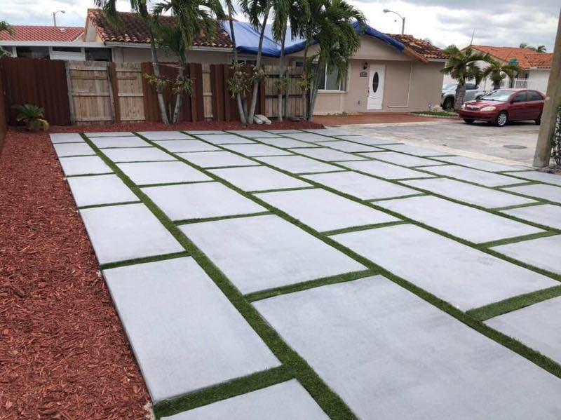 A modern concrete driveway with large rectangular slabs separated by strips of green grass. The decorative concrete driveway leads to a residential street with houses and a motorcycle parked in the distance. Bushes and trees line the edge of the driveway, showcasing Naples Concrete Solutions' craftsmanship.