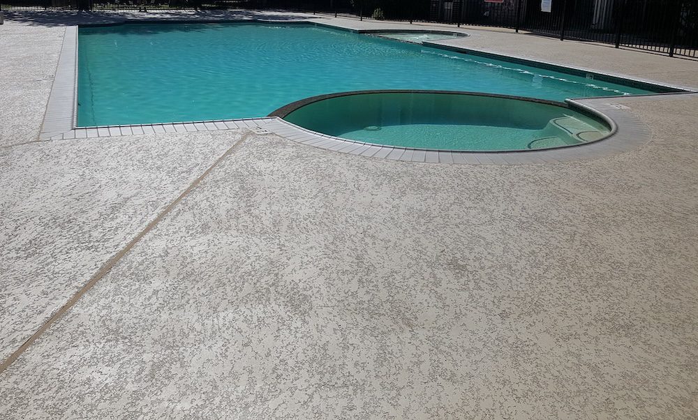 A view of a backyard swimming pool with clear blue water. The pool, crafted by experienced concrete contractors, has a curvy shape and is surrounded by a light-colored concrete deck. In the background, there are trees and grassy areas, with a small rock formation to the left.