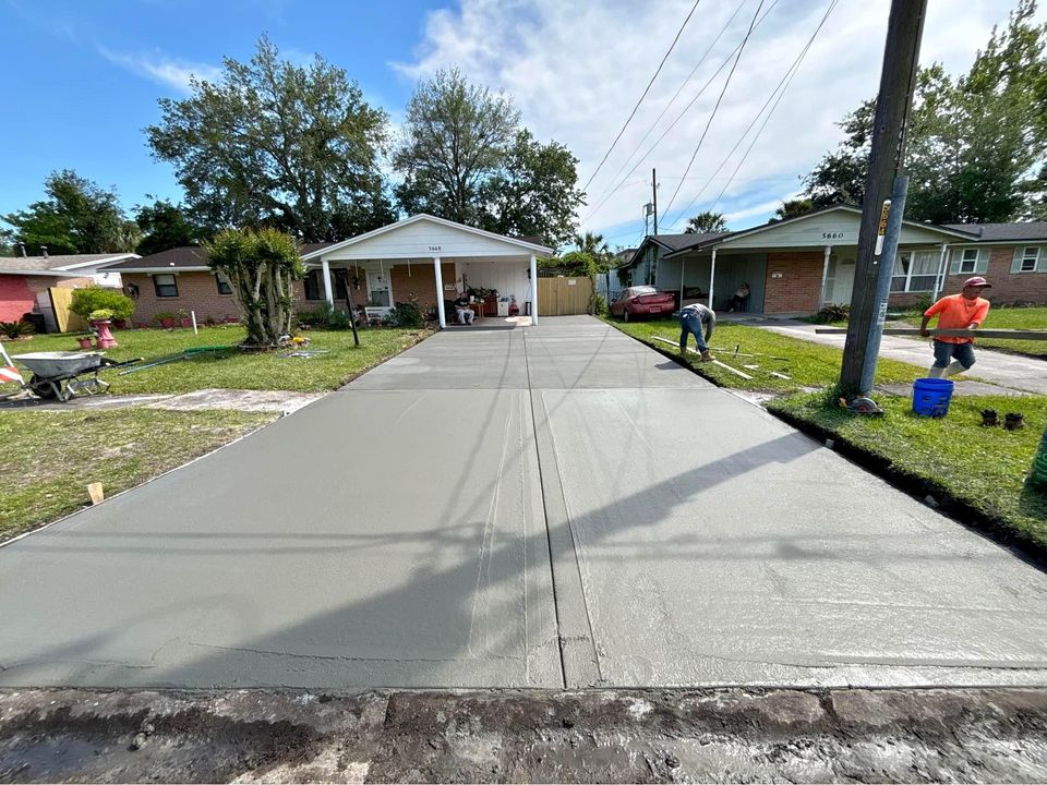 Two construction workers in fluorescent yellow shirts and work boots are standing on wet concrete. One operates a mechanical trowel, while the other smooths the surface of the new concrete driveway with a long-handled tool. A grassy area is visible in the background.