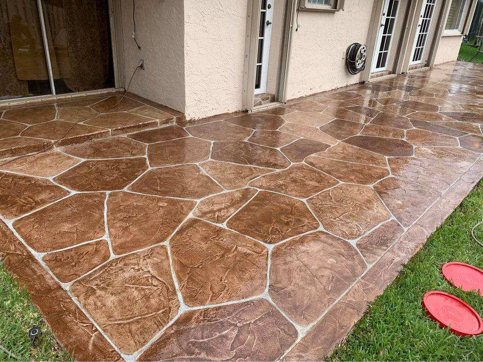 A backyard patio with a large, stamped concrete design featuring irregular, stone-like patterns in shades of brown. The patio adjoins a beige stucco house with multiple glass doors. A grass lawn borders the patio, where Naples Concrete Solutions recently completed resurfacing work, with two red frisbees lying on the ground.