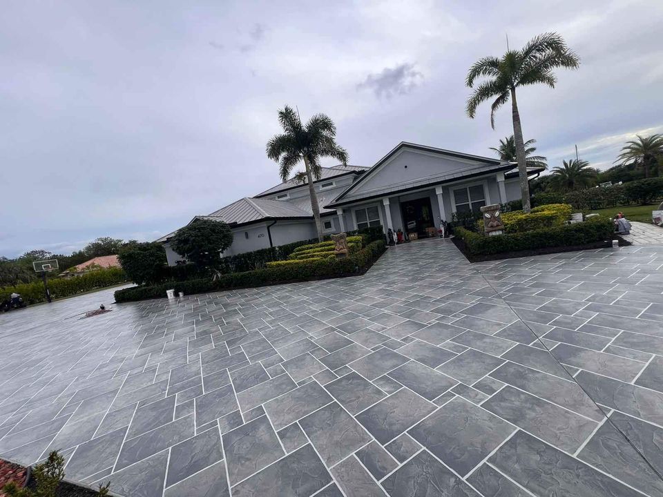 A large modern house with a gray tiled roof and white exterior walls sits at the end of a spacious, neatly tiled driveway created by experienced concrete contractors in Naples, FL. Palm trees and well-maintained hedges decorate the front yard. The sky is overcast, hinting at cloudy weather.