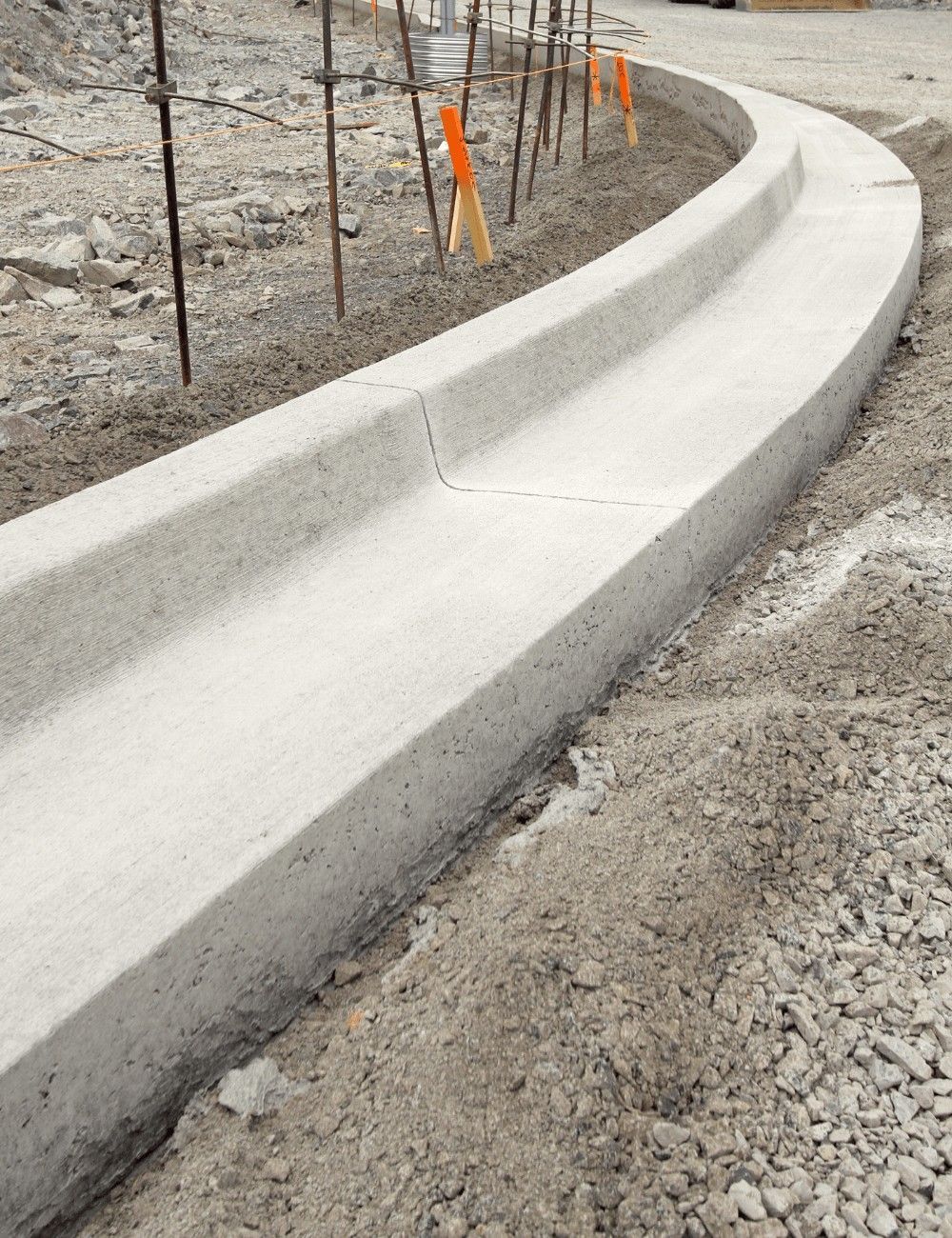 A freshly paved concrete curb on a construction site by Naples Concrete Solutions. The curb follows a curved path and is flanked by soil and gravel. Metal stakes with orange markers line the left side, indicating ongoing work. An ADA-compliant ramp is also being constructed for enhanced accessibility.