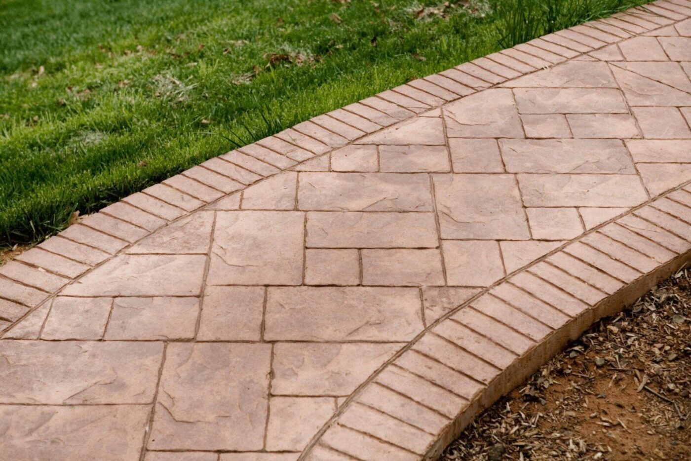 A paved stone walkway with a rectangular pattern is bordered by uniformly laid narrow bricks. The walkway, crafted by licensed decorative concrete contractors in Bonita Springs, FL, curves gently, separating a grassy lawn on the left from a patch of bare soil or mulch on the right.