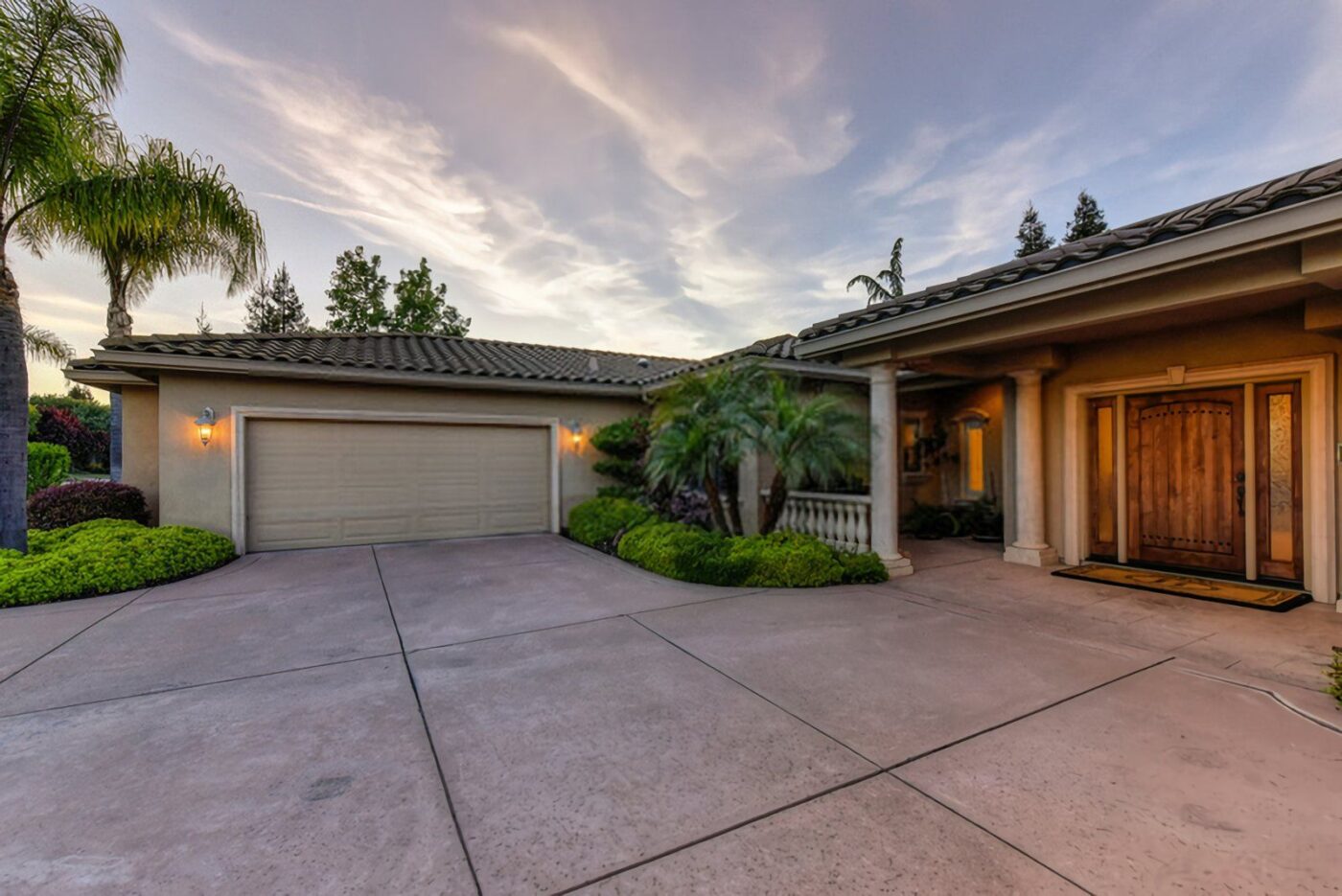 Wide-angle view of a single-story house with a green-tiled roof, double garage, and wooden front door in Golden Gate FL. The well-maintained property features a spacious driveway with decorative concrete, green shrubs, and palm trees against a backdrop of a sunset sky with soft clouds.