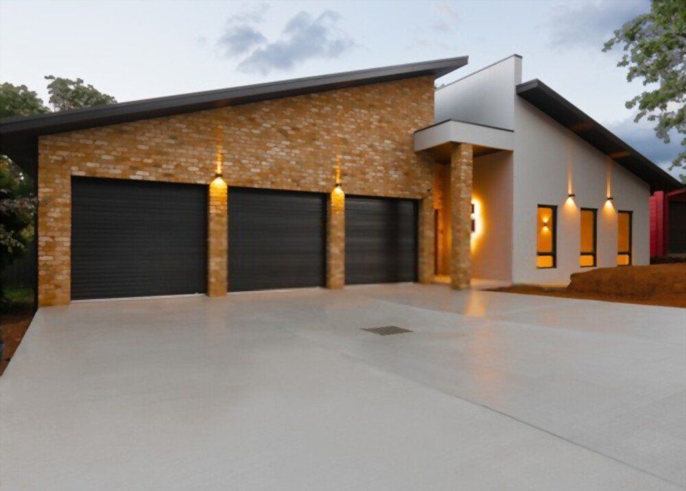 A modern house with a combination of brick and white exterior walls, featuring three black garage doors, warm outdoor lighting, a slanted roof, and a smooth concrete driveway crafted by Naples Concrete Solutions. The garage lights are turned on, and the sky is partly cloudy.