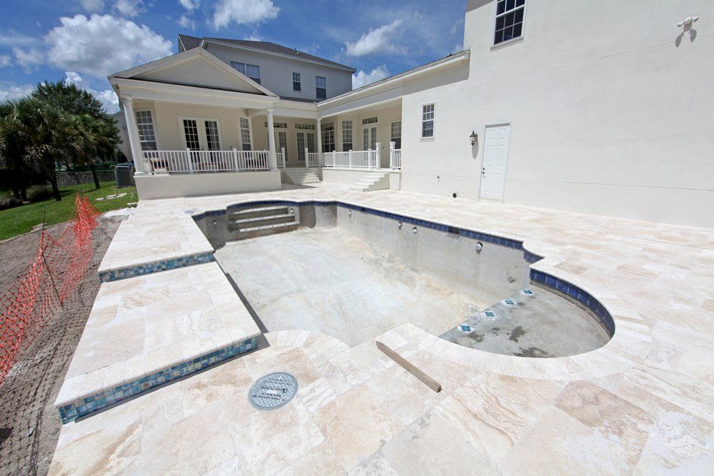 Unfinished swimming pool in a backyard with white tile surroundings. Adjacent to a large white house with multiple windows and a porch, partially cloudy sky in the background. Orange safety netting borders part of the area. Naples Concrete Solutions offers a free estimate for finishing your decorative pool deck.