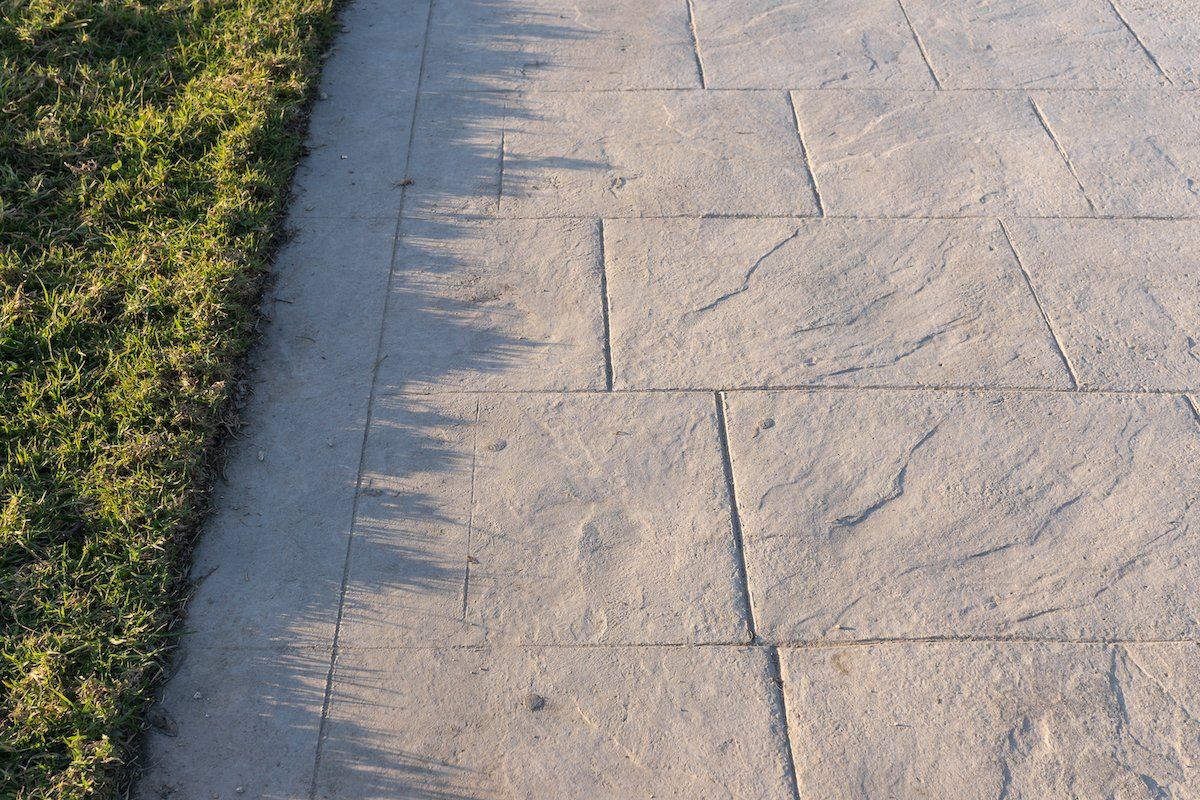 The image shows a segment of a paved walkway with large, rectangular tiles next to a grassy area. The walkway casts a shadow on the grass, creating a defined line between the grass and the pavement. The sunlight creates a clear contrast between the two surfaces, showcasing quality concrete work typical of Marco Island.