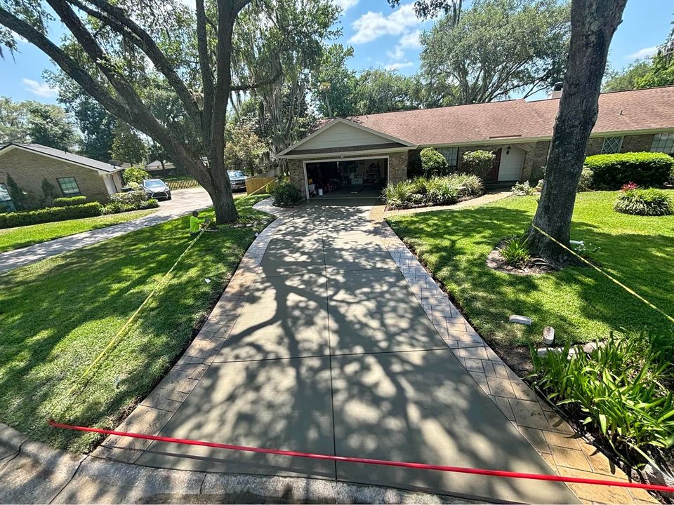 A freshly poured concrete driveway by Naples Concrete Solutions, featuring a red line marked near the entrance and yellow caution tape on either side, leads to a single-story house surrounded by lush, green landscaping and tall trees. The driveway is flanked by well-maintained grassy areas.